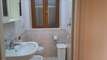 Une salle de bain avec un lavabo, deux toilettes, une fenêtre et une poubelle.