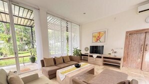 Un salon moderne comprenant un canapé, un fauteuil et un pouf, un téléviseur à écran plat et une grande fenêtre donnant sur un paysage verdoyant.