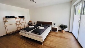 Ein modernes Schlafzimmer mit Holzboden, einem Bett mit schwarz-weißer Bettwäsche, einem Nachttisch mit Lampe, einem kleinen Hocker, einer Wandhalterung und einem Topfpflanzen.