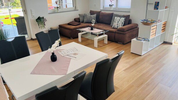 Ein modernes Wohnzimmer mit einem braunen Sofa, einem weißen Couchtisch und schwarzen Stühlen. Es gibt ein weißes Bücherregal und ein Fenster mit Jalousien.