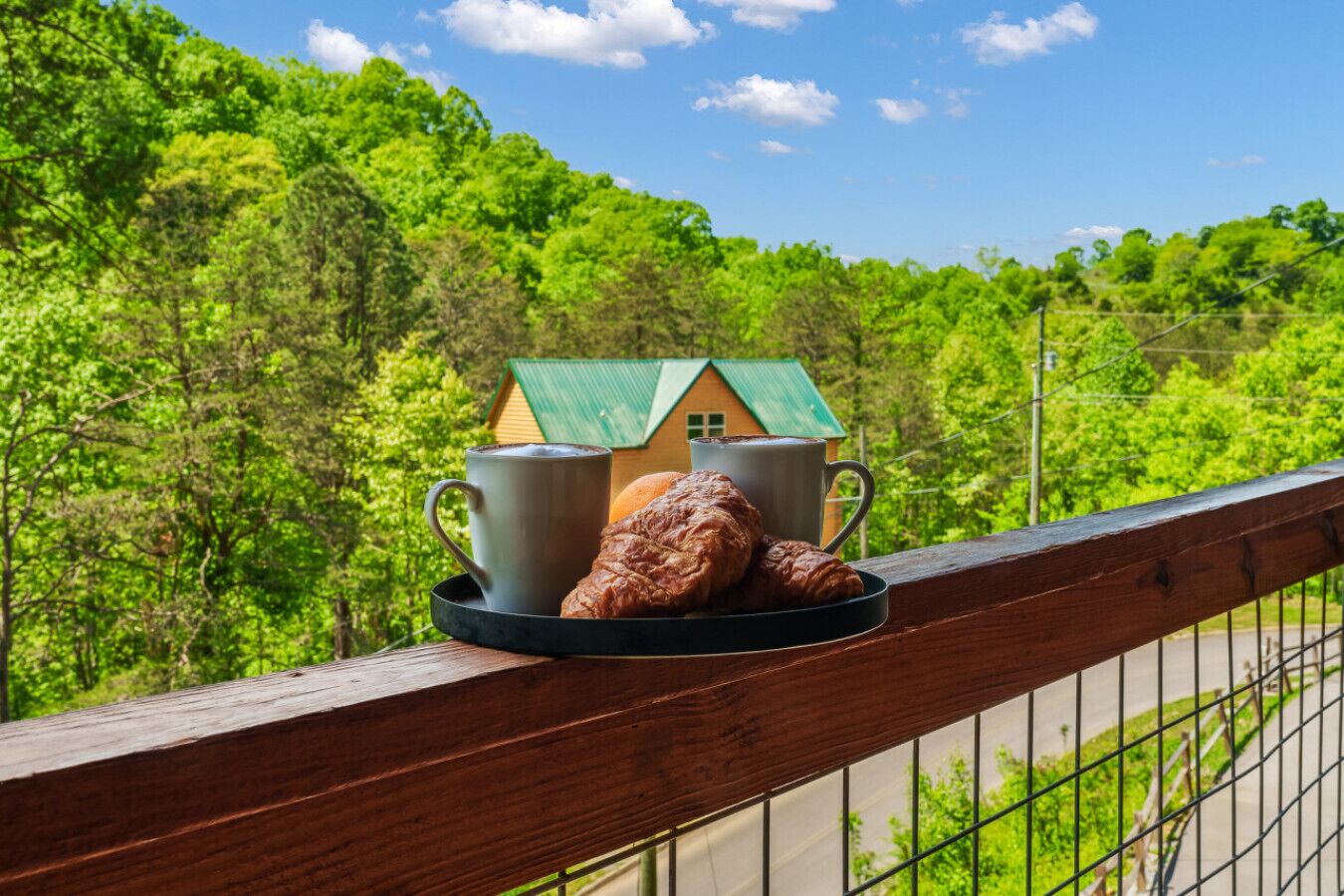 Un balcon avec un plateau de croissants et deux tasses, donnant sur une forêt verdoyante et une petite maison au toit vert.
