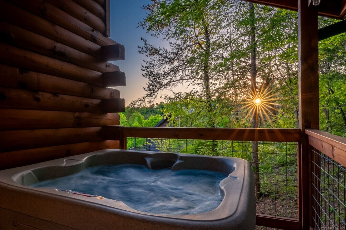 A hot tub on a wooden deck with a view of trees and a sunset.