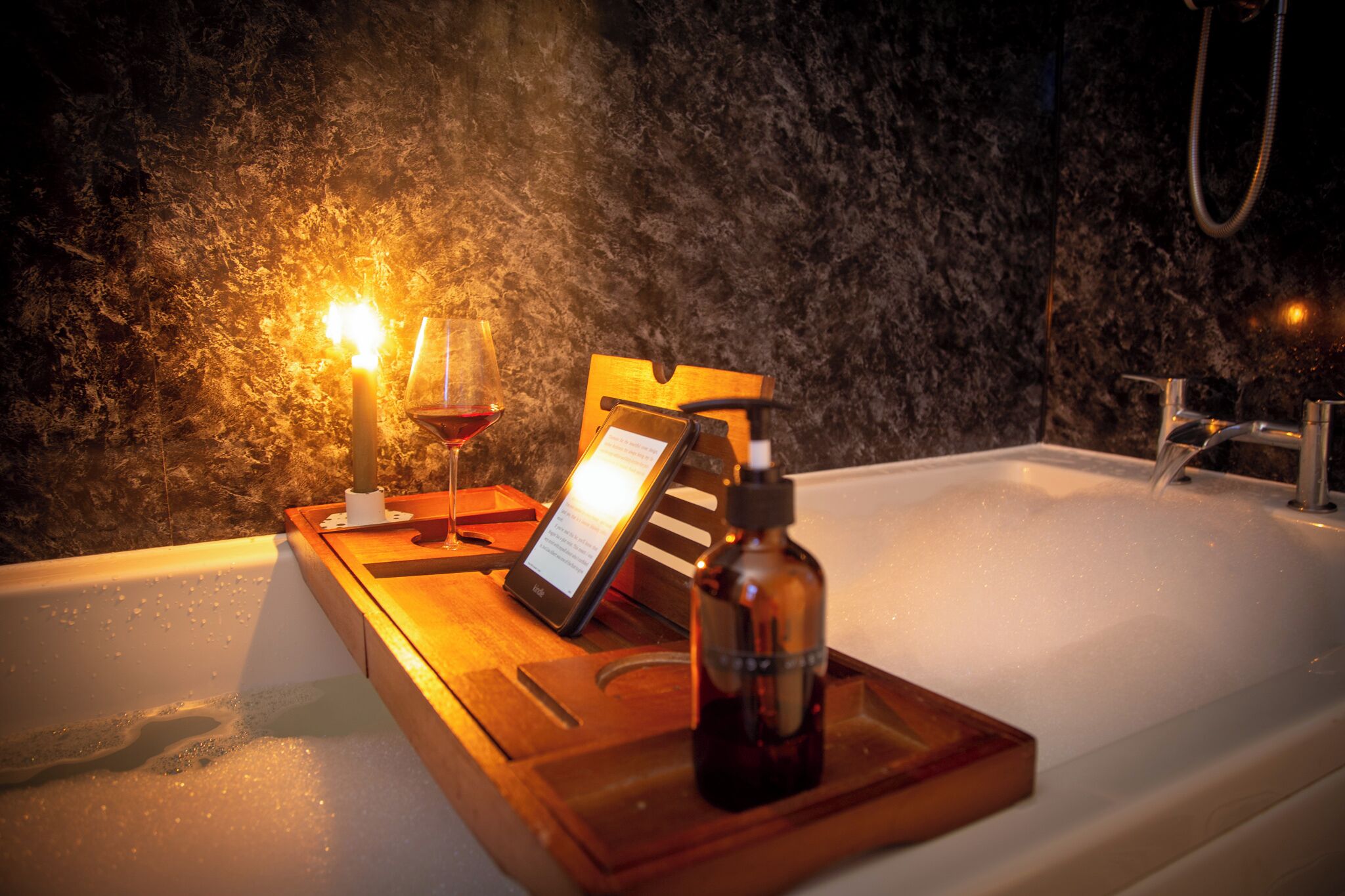 Une baignoire avec un plateau en bois sur lequel sont posés un flacon et une tablette, le tout placé devant un mur à texture.