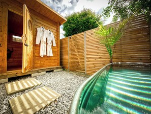 Un edificio de spa de madera con puerta abierta, una piscina pequeña y una cerca de madera.