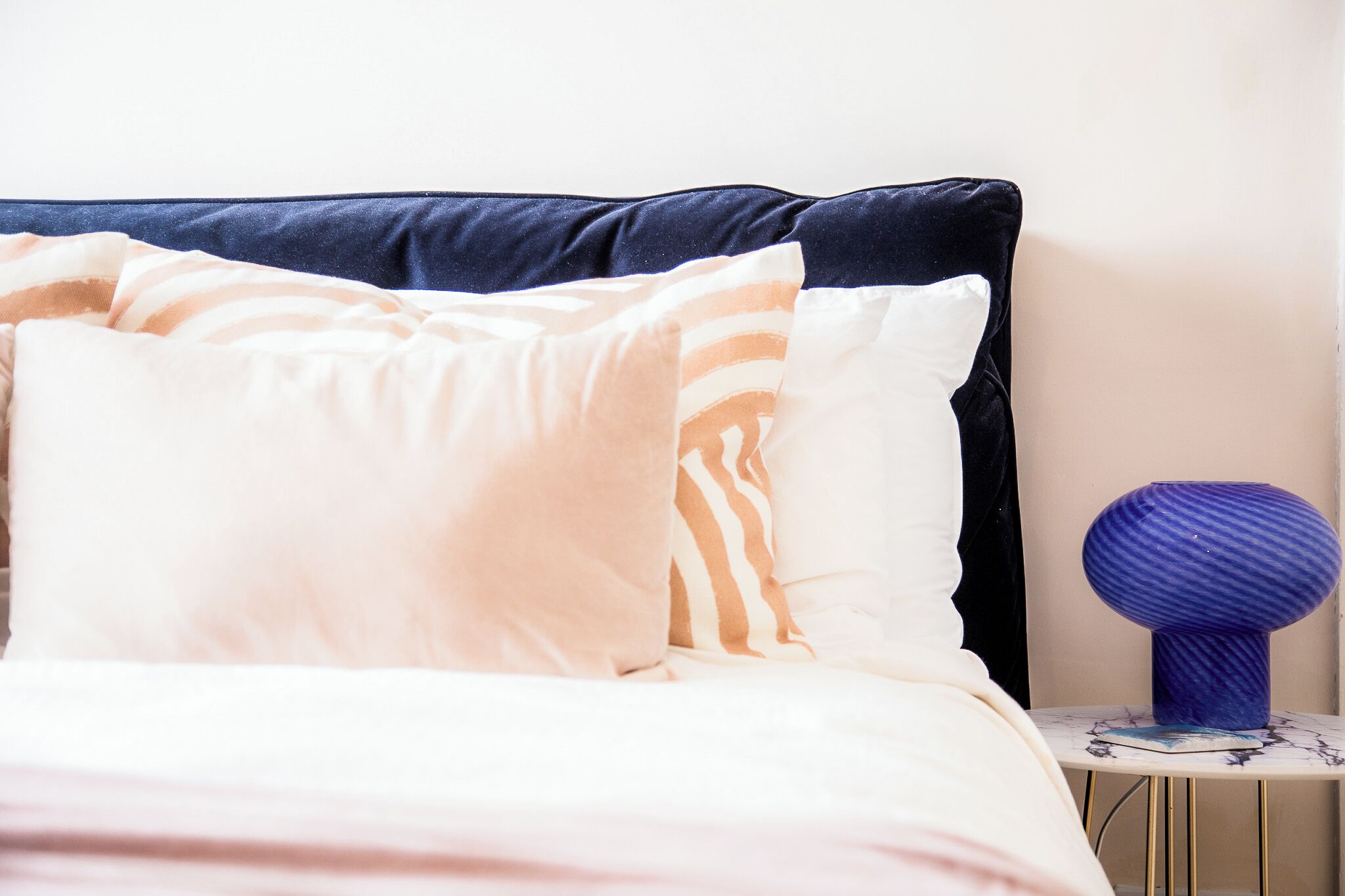 A bed with a dark headboard, a white and peach striped pillow, and a blue decorative object on a small table beside the bed.