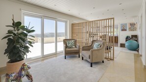 A spacious room with a large sliding glass door leading to a balcony, two armchairs with decorative pillows, a large potted plant, and a blue exercise ball. - Coastline Arrabida Villa by Cadenzalux (Sesimbra)