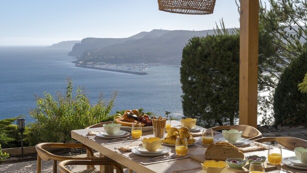 A breakfast table set with plates, glasses, and a variety of food items, overlooking a coastal landscape with cliffs. - Coastline Arrabida Villa by Cadenzalux (Sesimbra)