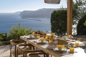 A breakfast table set with plates, glasses, and a variety of food items, overlooking a coastal landscape with cliffs. - Coastline Arrabida Villa by Cadenzalux (Sesimbra)