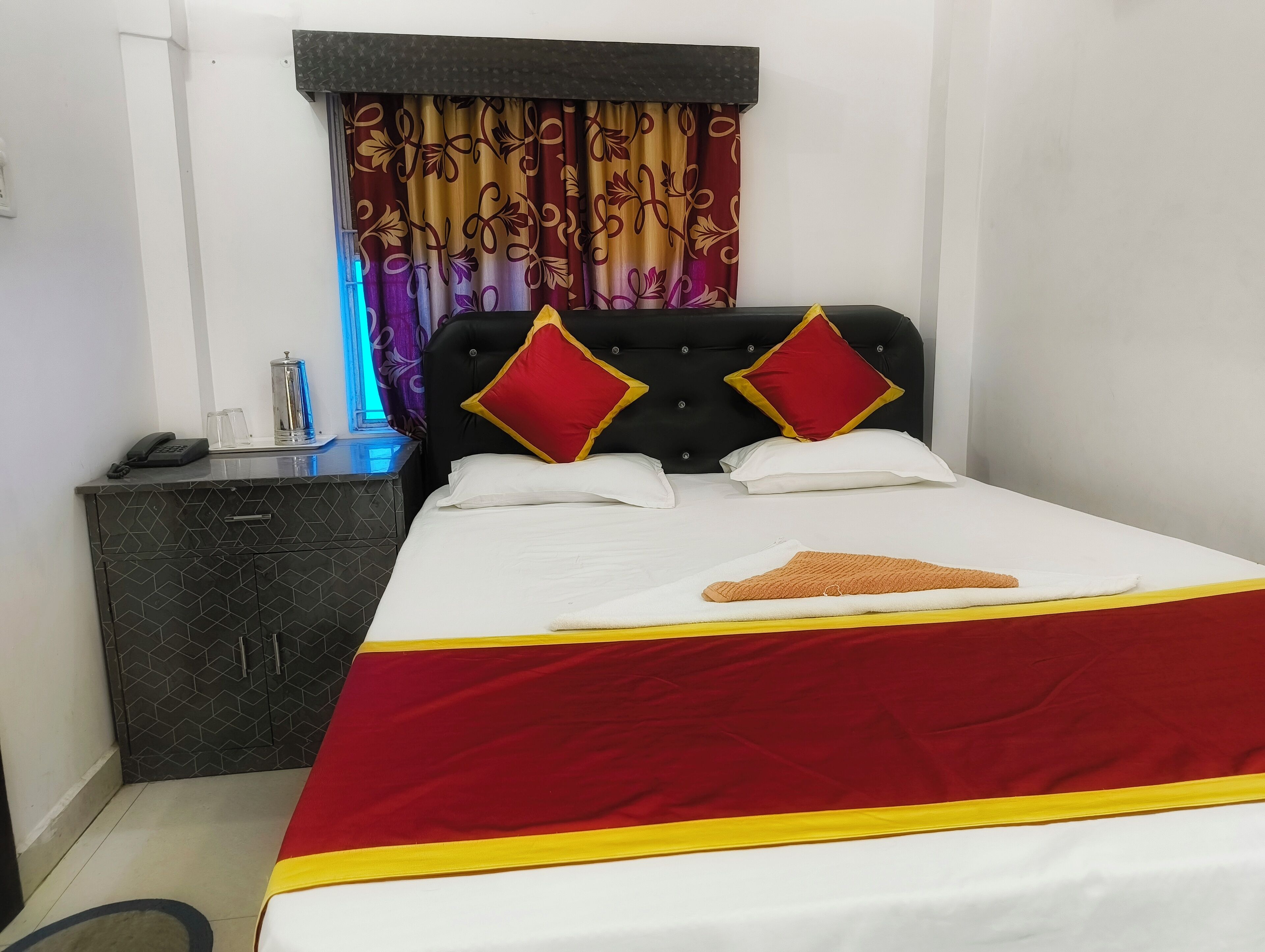 A neatly made bed with a red and yellow bedspread, two red and yellow pillows, and a single white pillow. A small bedside table with a phone and a glass of water is visible.