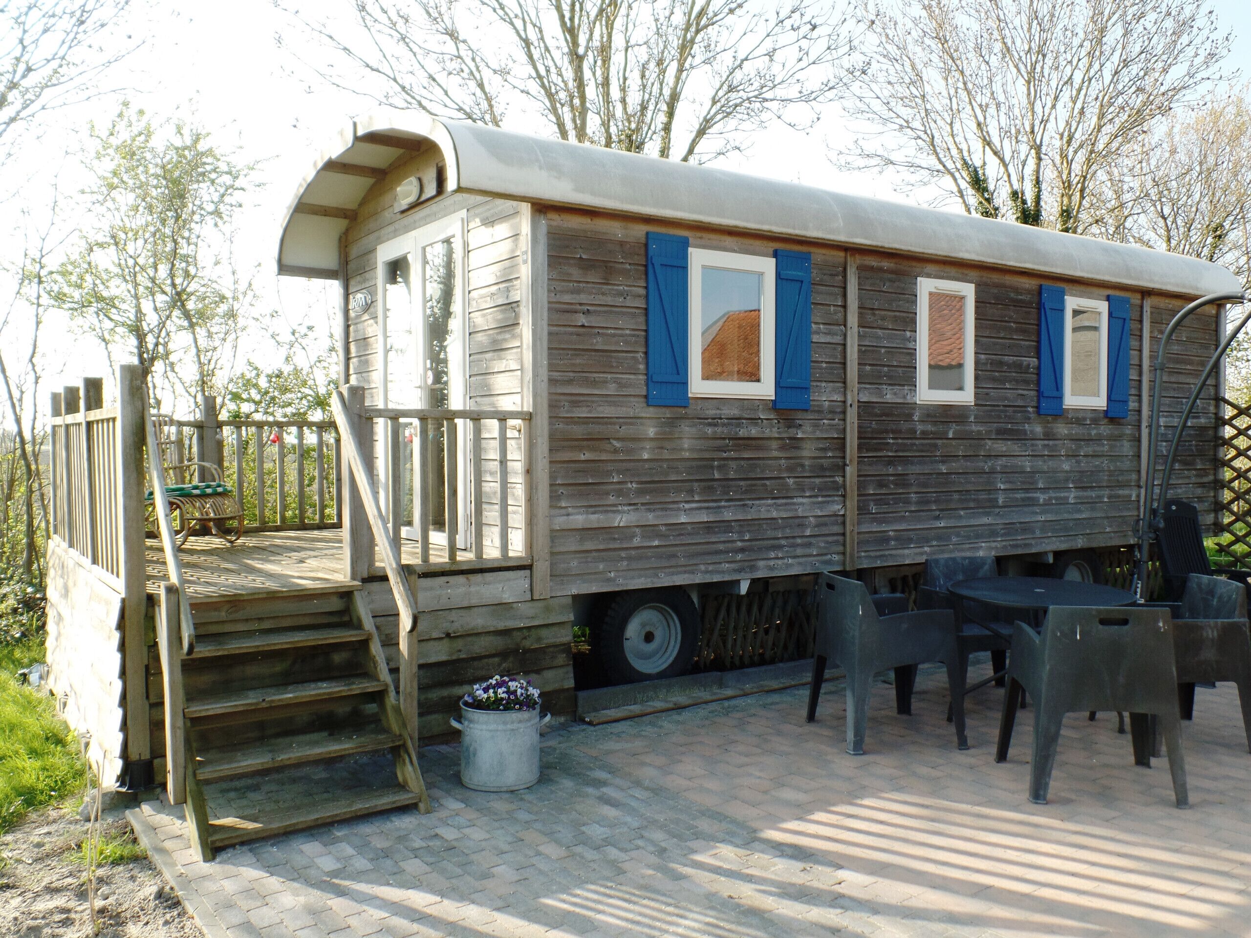 Een houten treinwagon met een overdekte veranda en een tafel met stoelen er buiten.