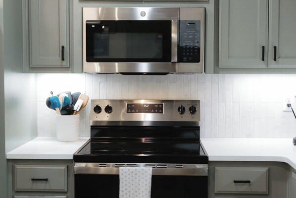 Une cuisine équipée d’un four à micro-ondes, d’un four et d’une cuisinière en acier inoxydable. On y trouve des placards au-dessus de la cuisinière et un pare-feu en carreaux blancs.