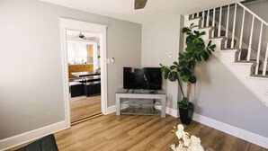 A living room with a television on a glass stand, a ceiling fan, a staircase, and a potted plant.