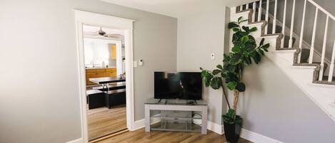 A living room with a television on a glass stand, a ceiling fan, a staircase, and a potted plant.