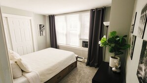 A bedroom with a bed, a window with curtains, a radiator, a plant, and wall art.