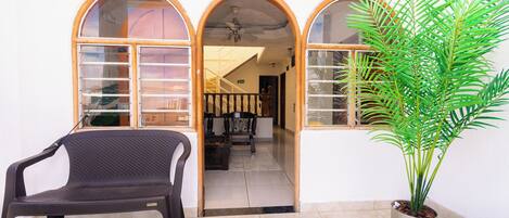 A room with a dark plastic chair, a potted plant, and a view through an arched doorway to a staircase and a living area.