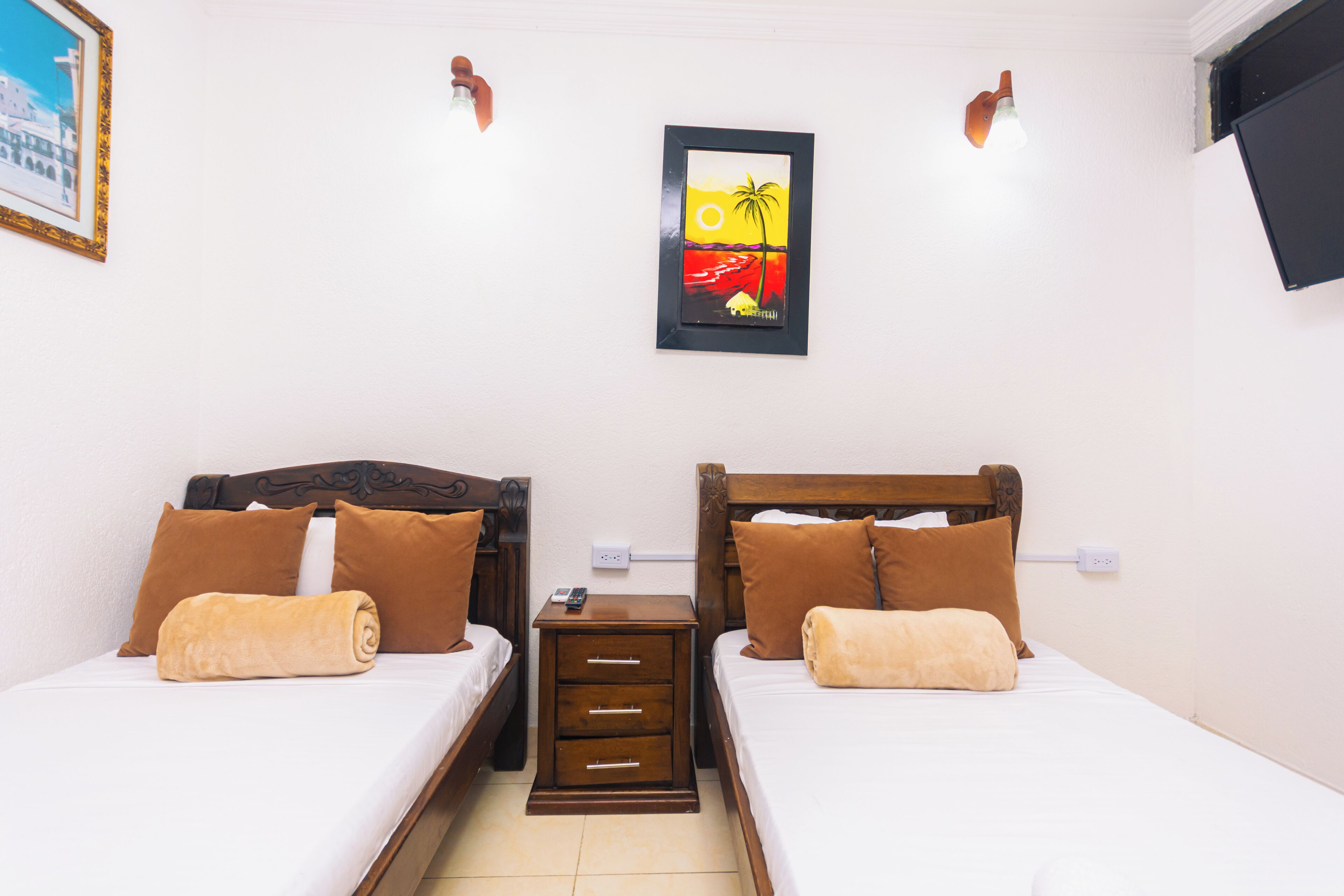 A hotel room with two beds, brown pillows, a nightstand, a framed picture, and a wall-mounted light fixture.