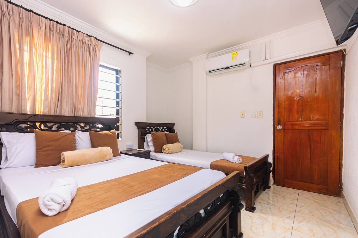 Habitación de hotel con dos camas, un armario de madera, una ventana con cortinas, un equipo de aire acondicionado y una puerta de madera.