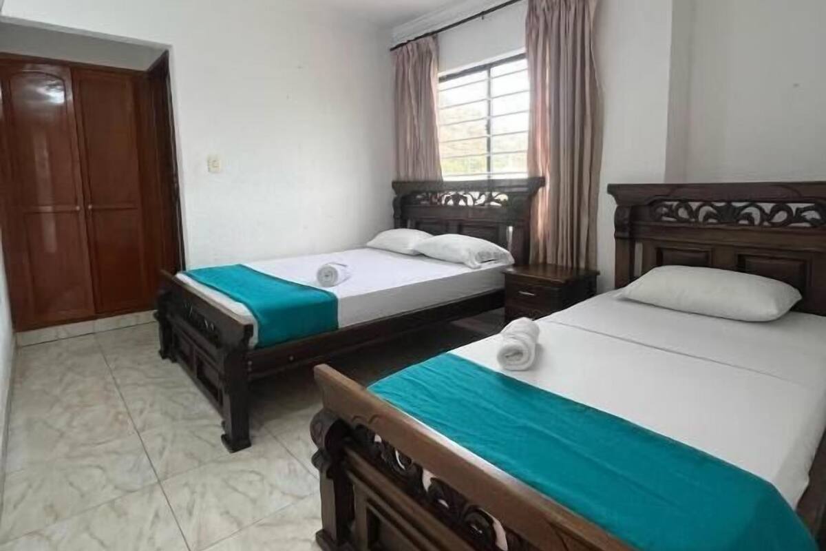 Una habitación de hotel con dos camas de madera, un ventanal con cortinas, un armario de madera y piso de baldosas.