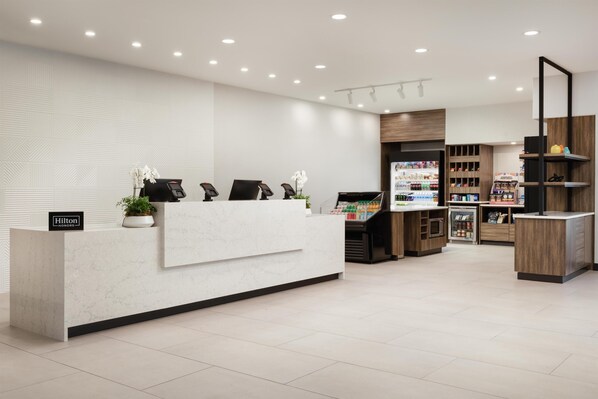 Ein moderner Hotel-Empfangsbereich mit Marmor-Theke, einem digitalen Bildschirm und einer Blumenkomposition. Mehrere Sitzbereiche mit Stühlen und einer kleinen Pflanze laden zum Verweilen ein. Die Rezeption wird von Regalen mit verschiedenen Gegenständen und einer Vitrine flankiert.