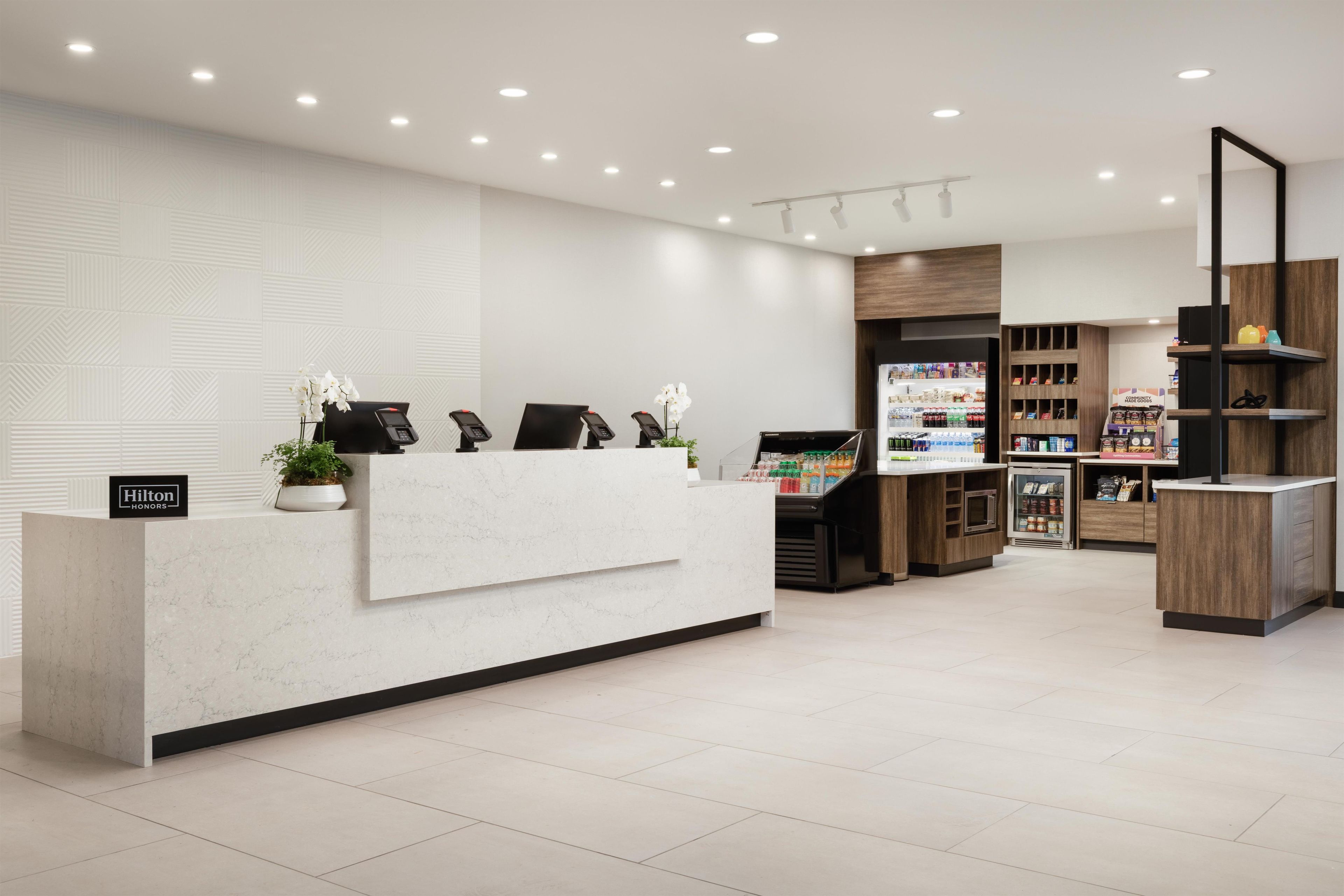 Un espace d’accueil moderne, avec un comptoir en marbre, un écran numérique et une composition florale. On y trouve plusieurs coins salon avec des chaises et une petite plante. Le comptoir d’accueil est bordé d’étagères où sont exposés divers objets, ainsi que d’une vitrine.