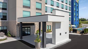 Ein moderner Hotelzugang mit überdachter Wartezone, Außenbestuhlung und Poolbereich im Hintergrund.
