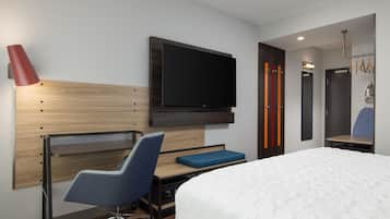A modern hotel room with a bed, desk, chair, TV, and a mirror.