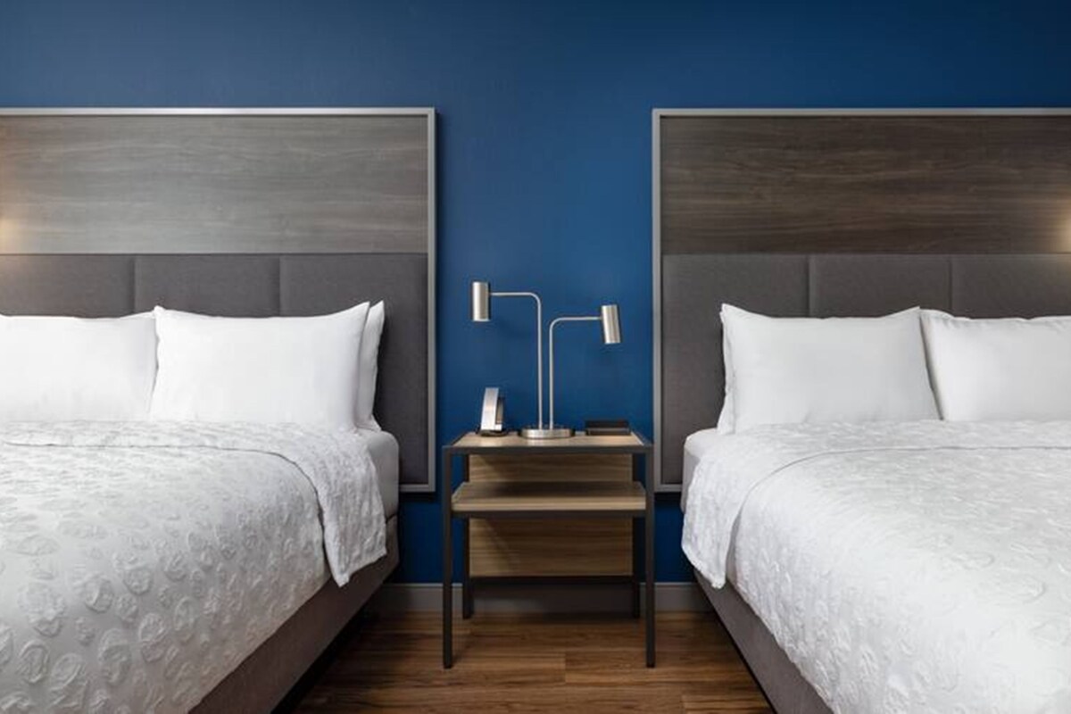 Habitación de hotel con dos camas, una mesita de noche con lámparas y una pared decorada en azul.
