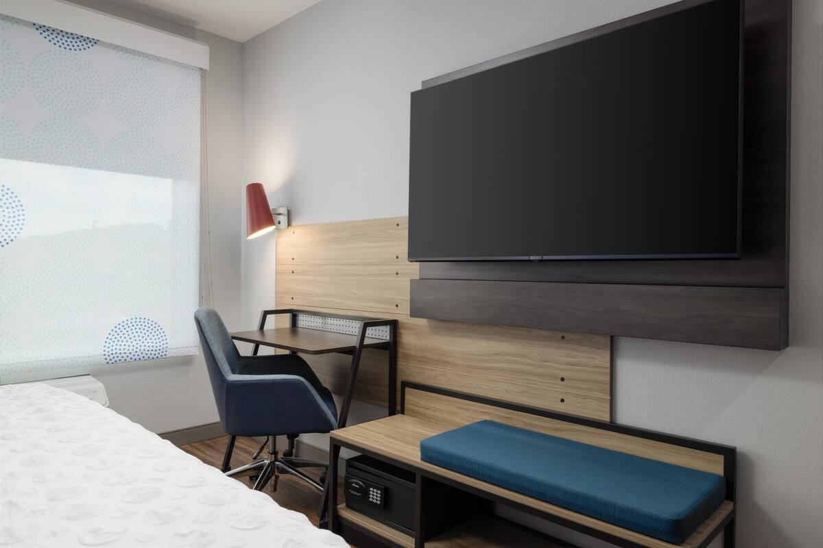 Una habitación de hotel moderna con cama, escritorio, silla, televisión y ventana con persianas.