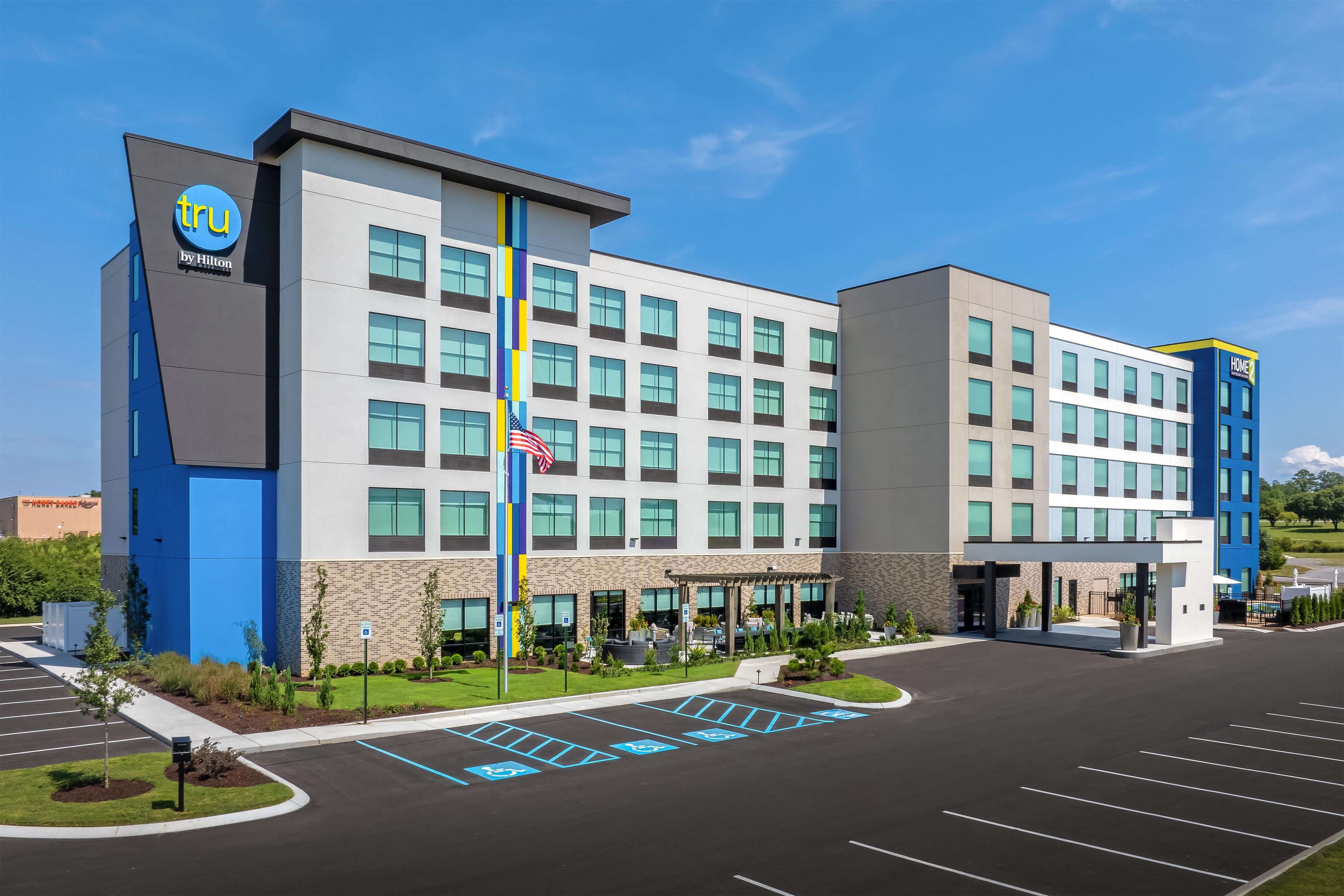 A modern hotel with a prominent 'tru by Hilton' sign, a parking lot with handicapped spaces, and a flagpole with an American flag.