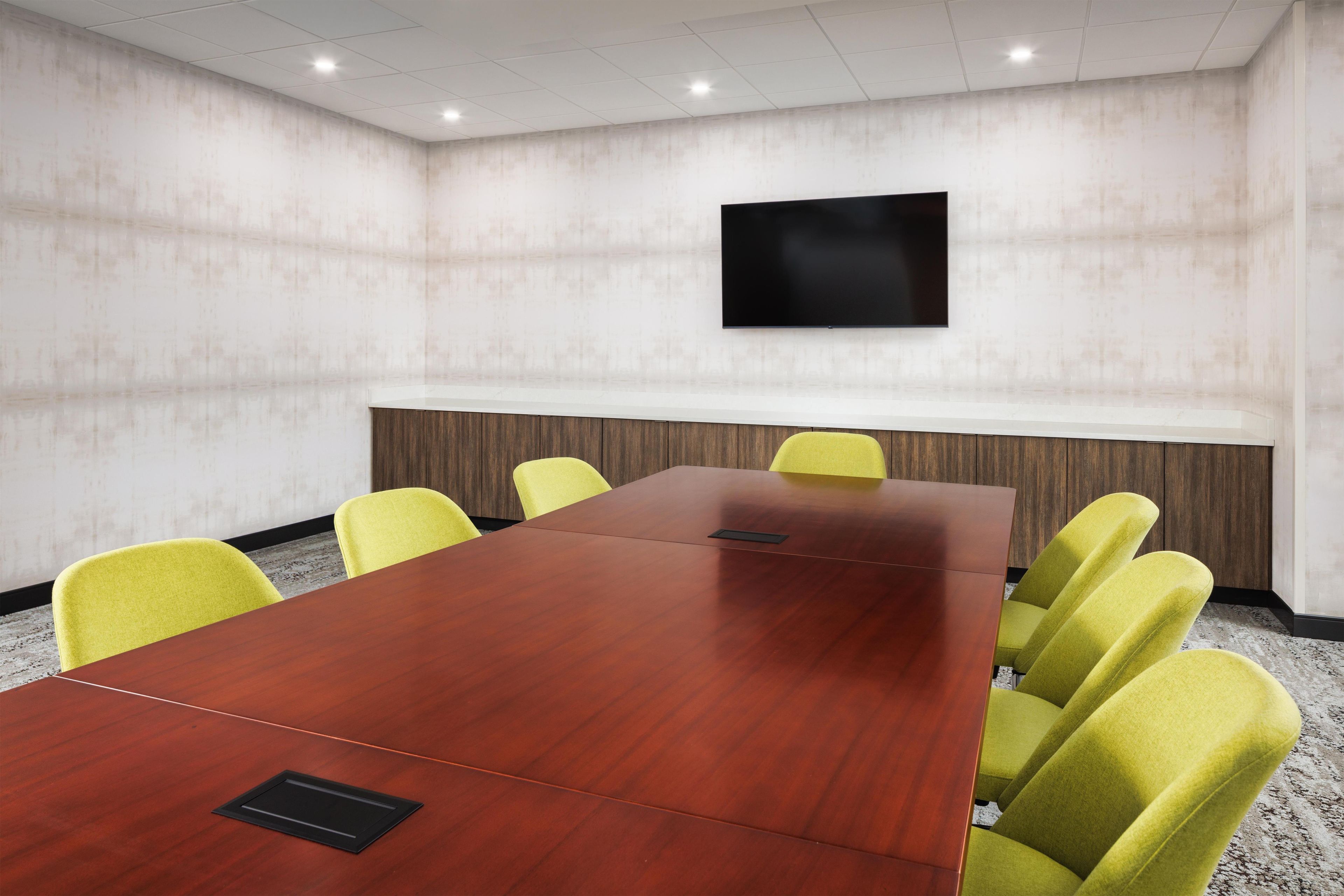 A conference room with a long wooden table, yellow chairs, a flat-screen TV mounted on the wall, and a whiteboard.