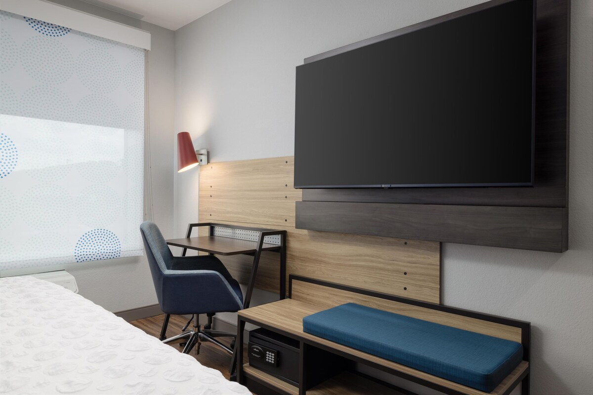 A modern hotel room with a bed, a desk, a chair, a TV, and a window with blinds.