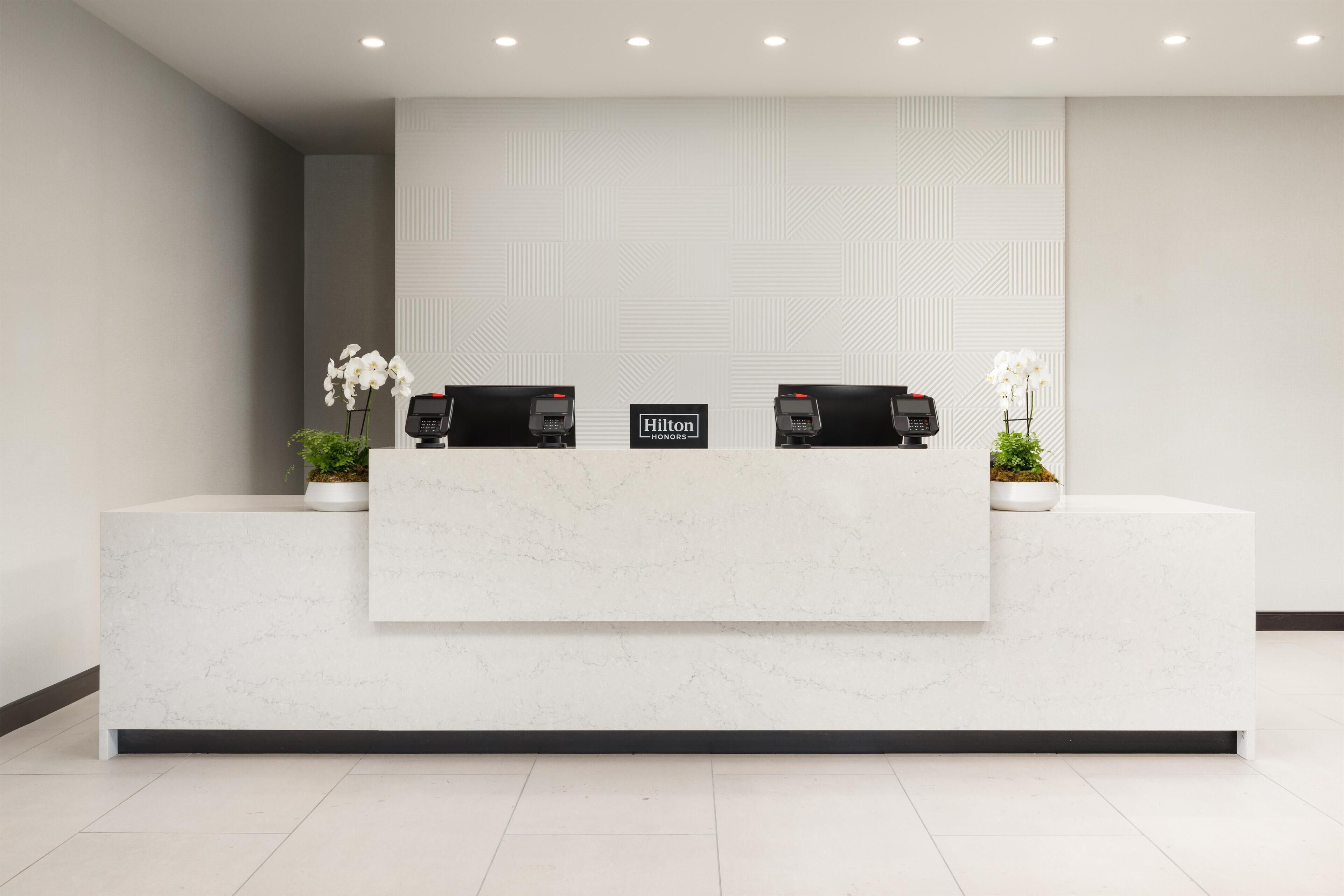 A clean, modern reception desk with two phones, a sign reading "Hilton Honors," and potted plants.