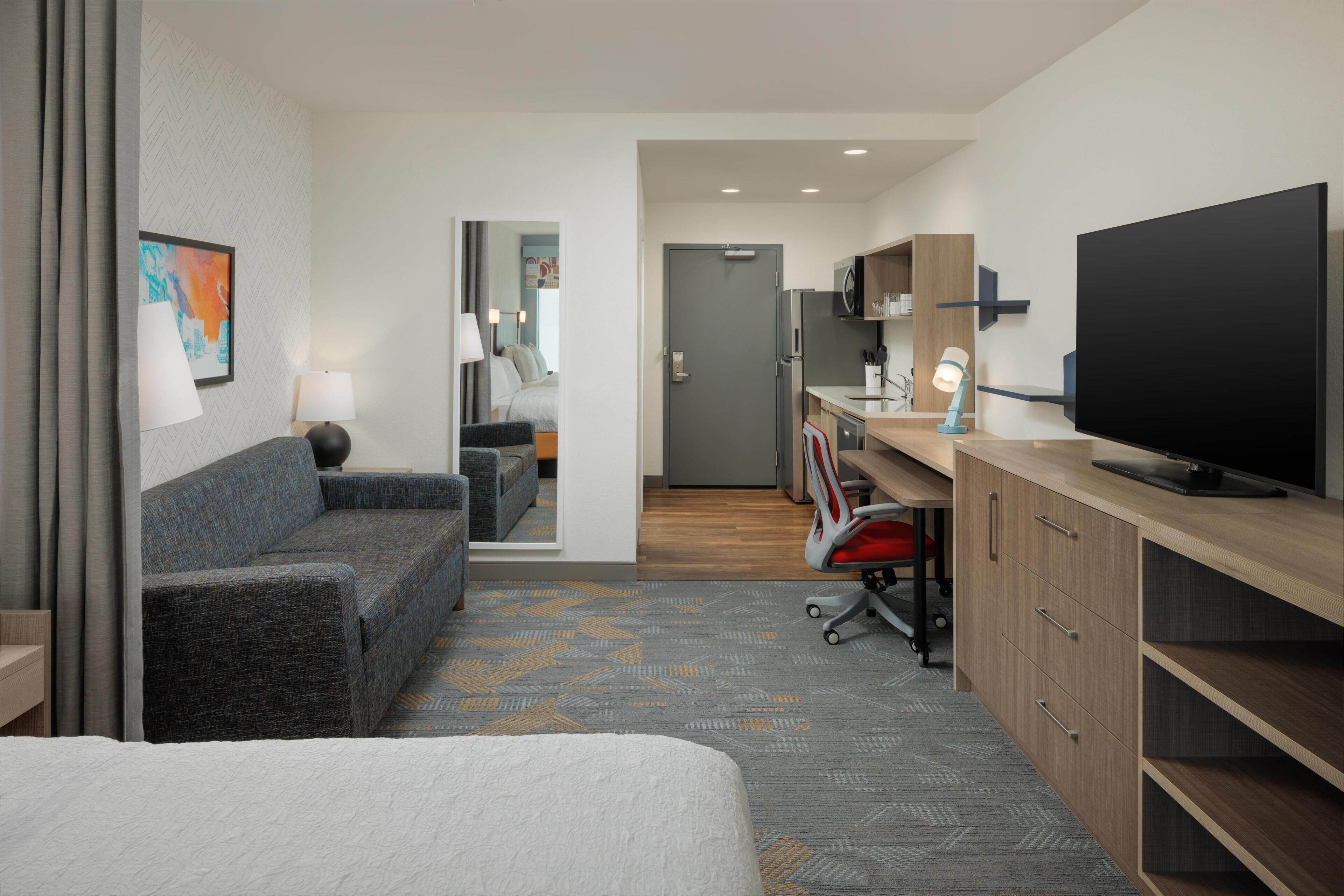 A hotel room with a bed, a sofa, a desk with a computer, a TV, and a kitchenette.