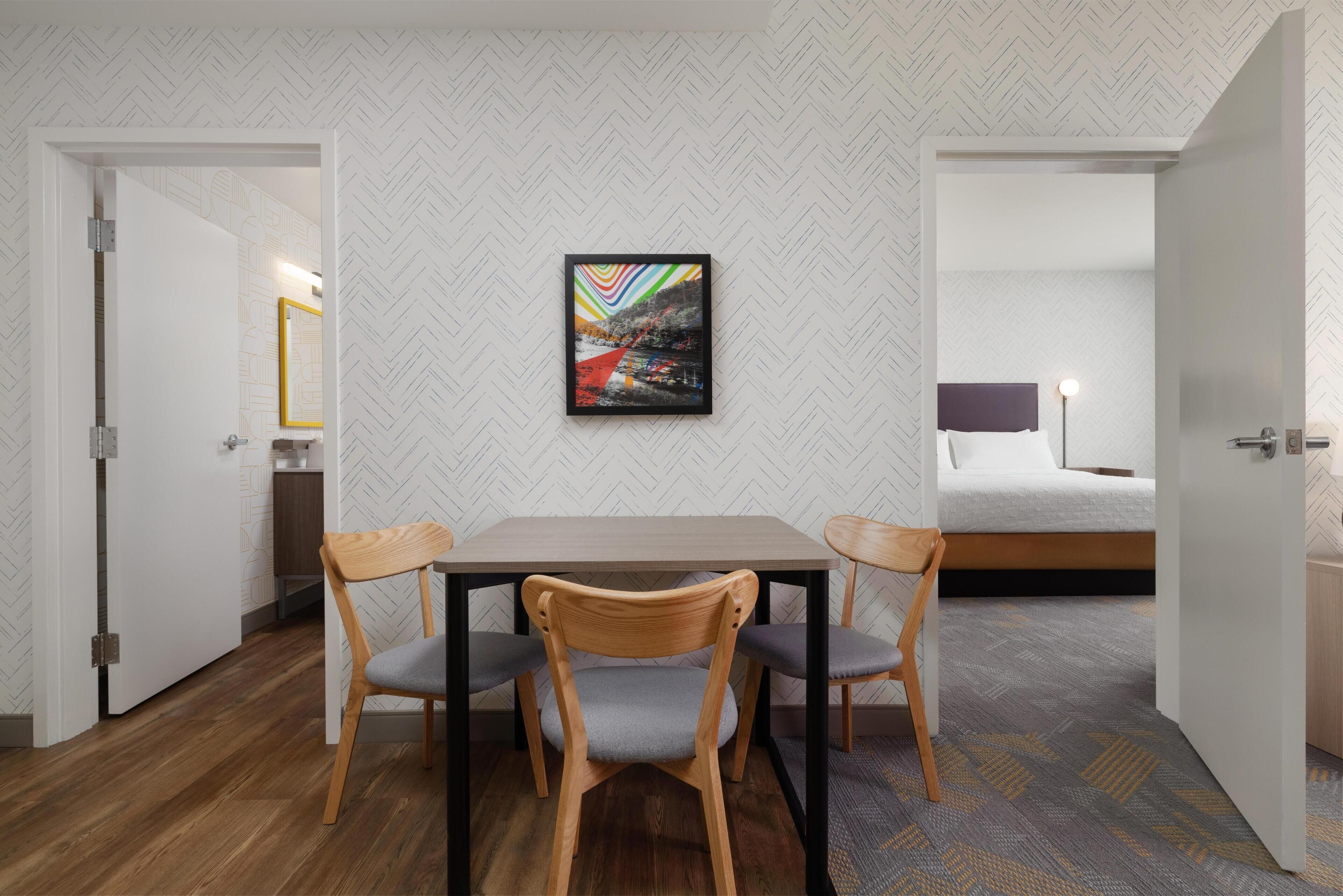 A hotel room with a wooden table and two chairs, a bed with a white headboard, and a framed picture on the wall.
