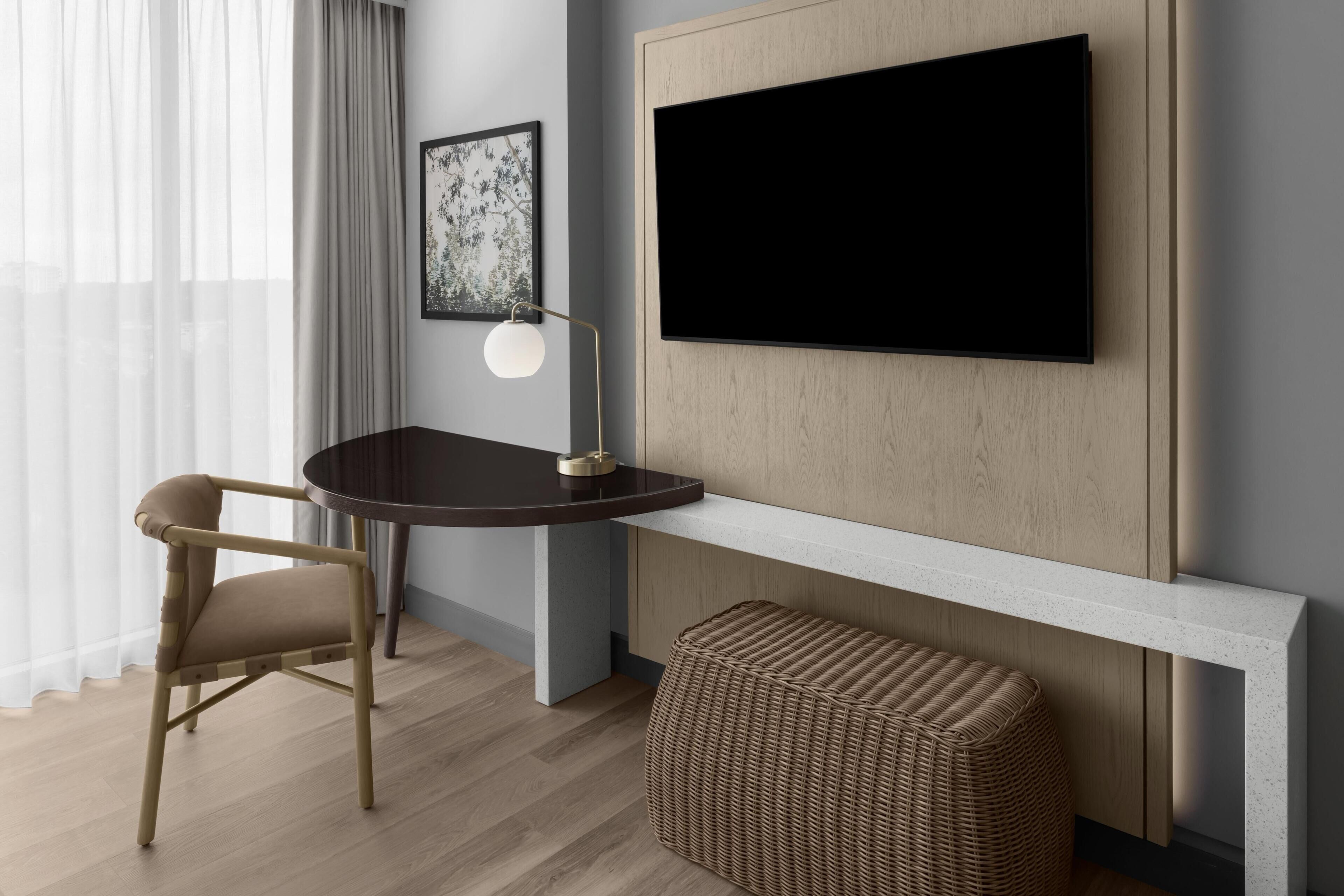Een moderne hotelkamer met een flatscreen televisie bevestigd aan een houten paneel, een ronde eettafel met een stoel, een vloerlamp en een geweven poef.