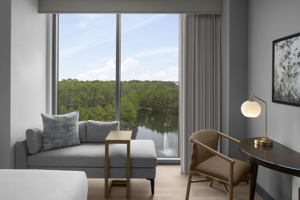 A modern hotel room with a large window offering a view of a forest and lake.