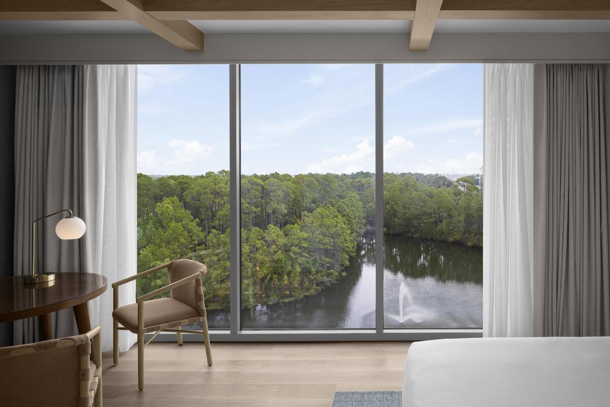 A modern hotel room with a large window offering a view of a forested area and a river. There is a bed, a chair, and a small table with a lamp.