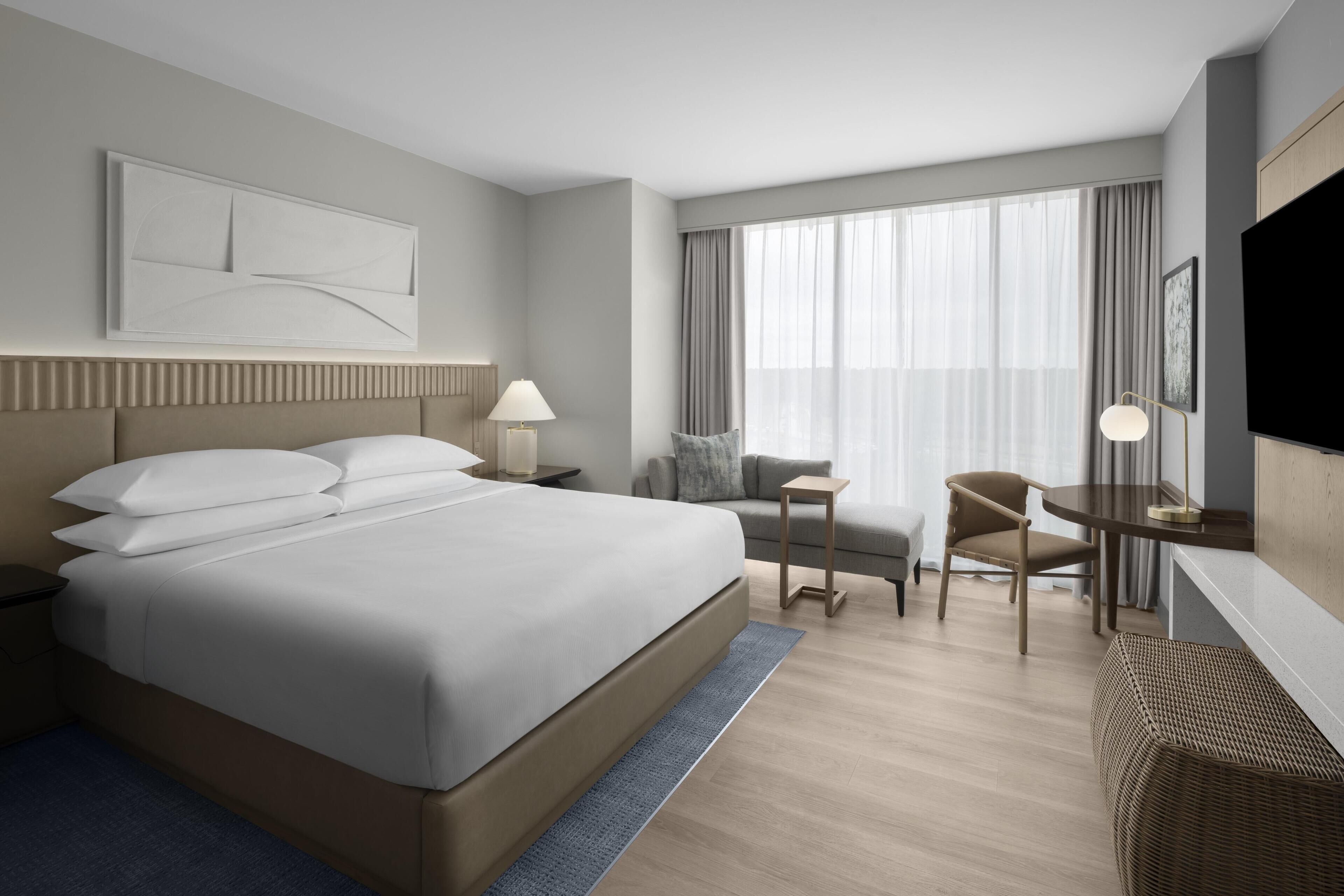 A modern hotel room with a large bed, a desk, a chair, a TV, and a window with curtains.