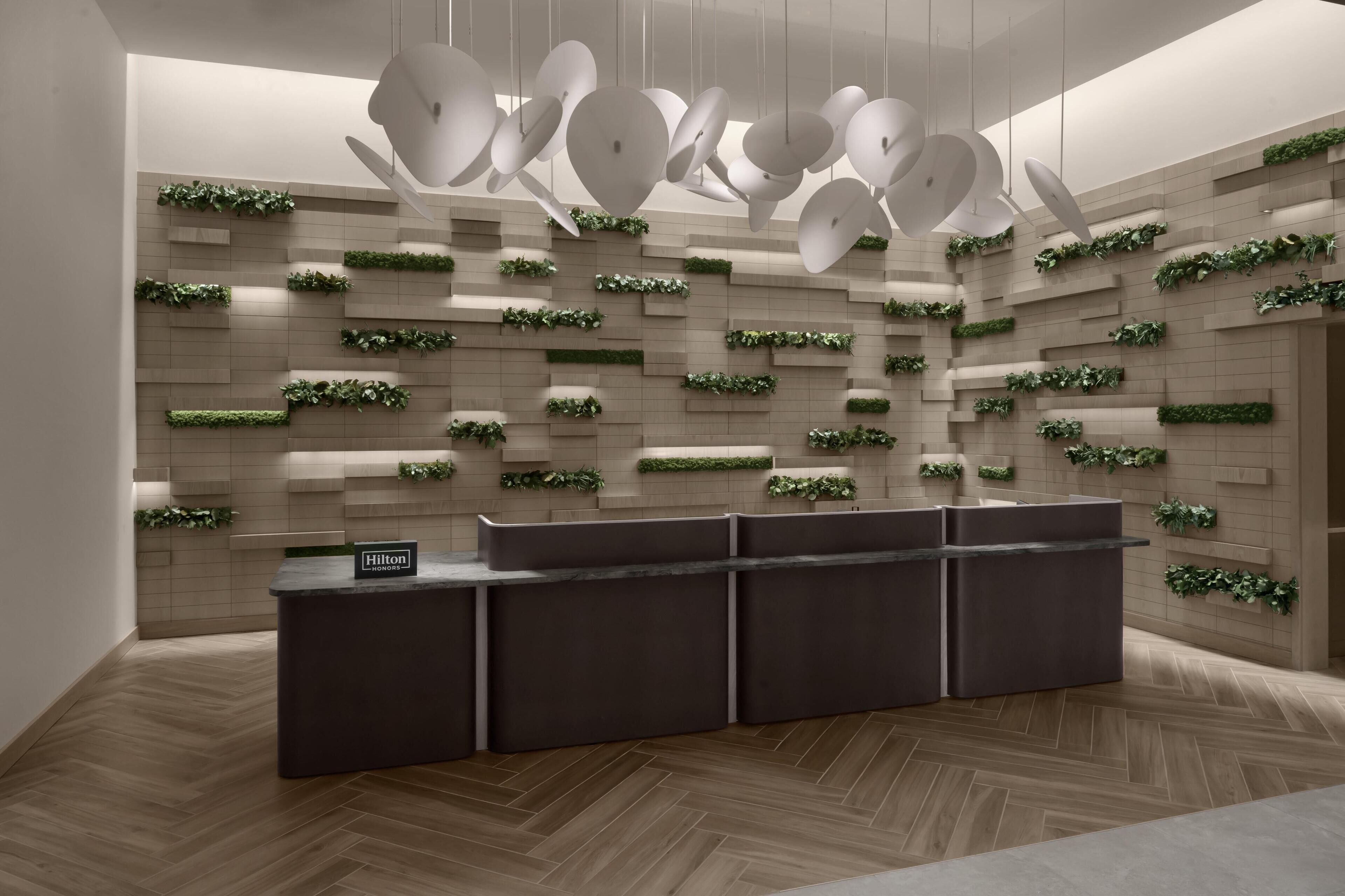 A modern hotel reception area with a dark reception desk, a wall-mounted plant display, and a unique ceiling light fixture.