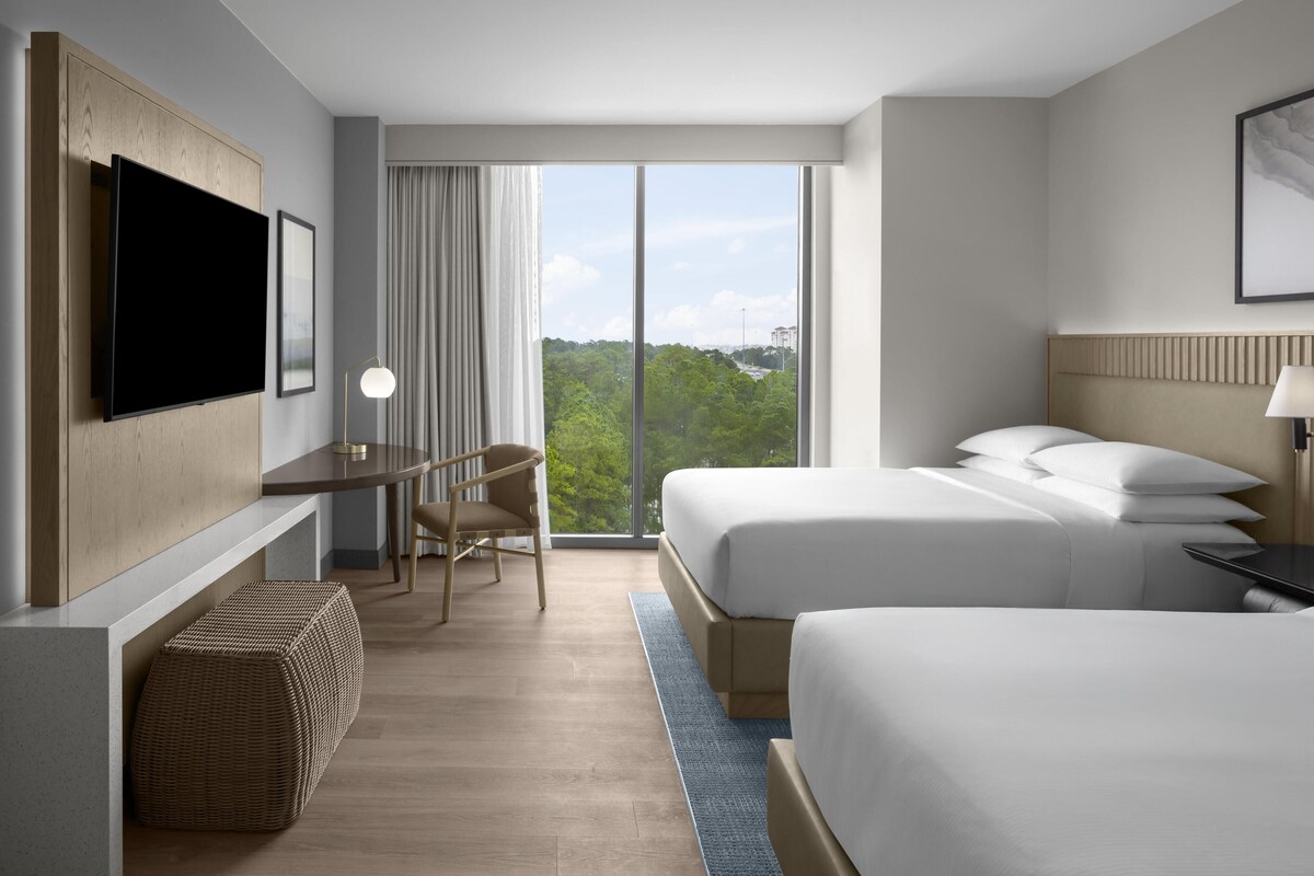 A modern hotel room with two beds, a desk, a flat-screen TV, and a large window offering a city view.