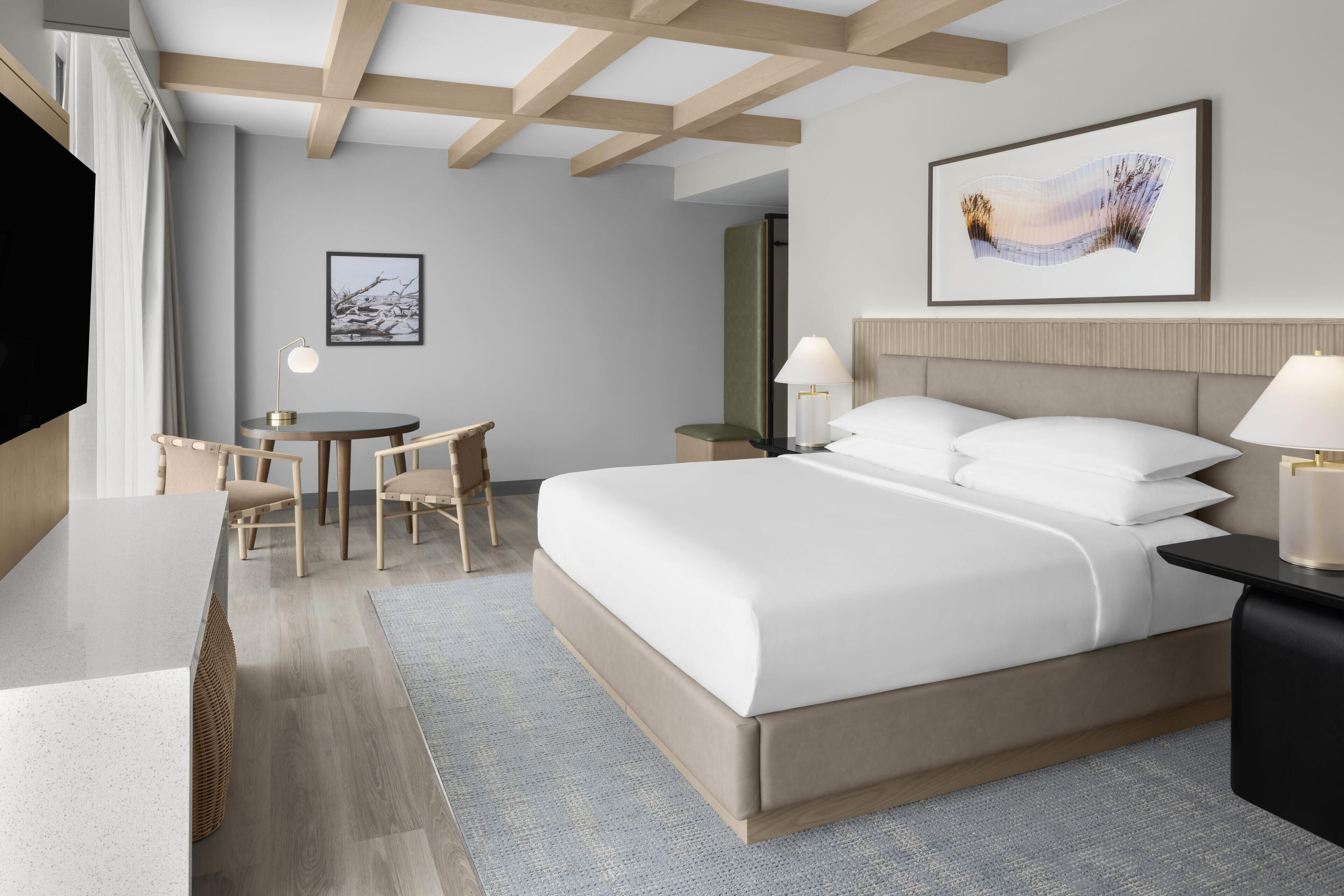 A modern hotel room with a large bed, a desk, and chairs. There is a flat-screen TV on the left and a framed picture on the wall.