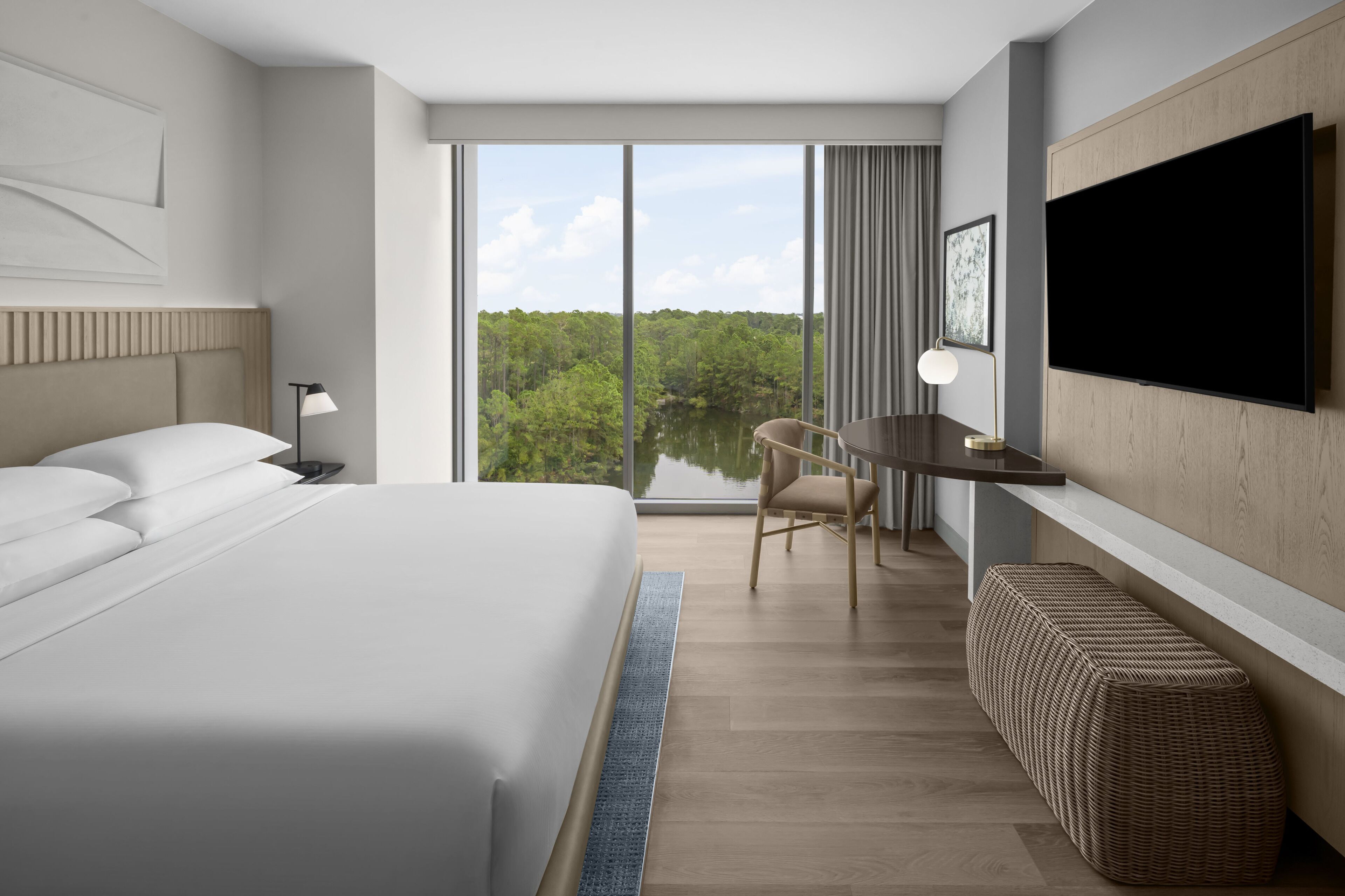 A modern hotel room with a large bed, a desk, a chair, a television, and a view of a lake.