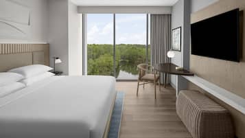 A modern hotel room with a large bed, a desk, a chair, a television, and a view of a lake.