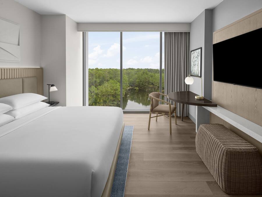 A modern hotel room with a large bed, a desk, a chair, a television, and a view of a lake.