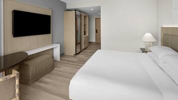 A hotel room with a bed, a TV mounted on the wall, a bedside table, a lamp, a wardrobe, and a small seating area with a bench.