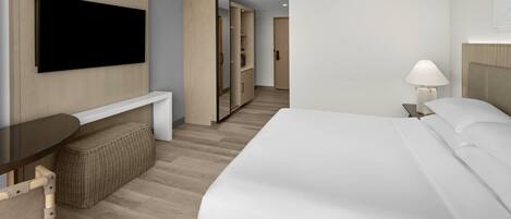 A hotel room with a bed, a TV mounted on the wall, a bedside table, a lamp, a wardrobe, and a small seating area with a bench.