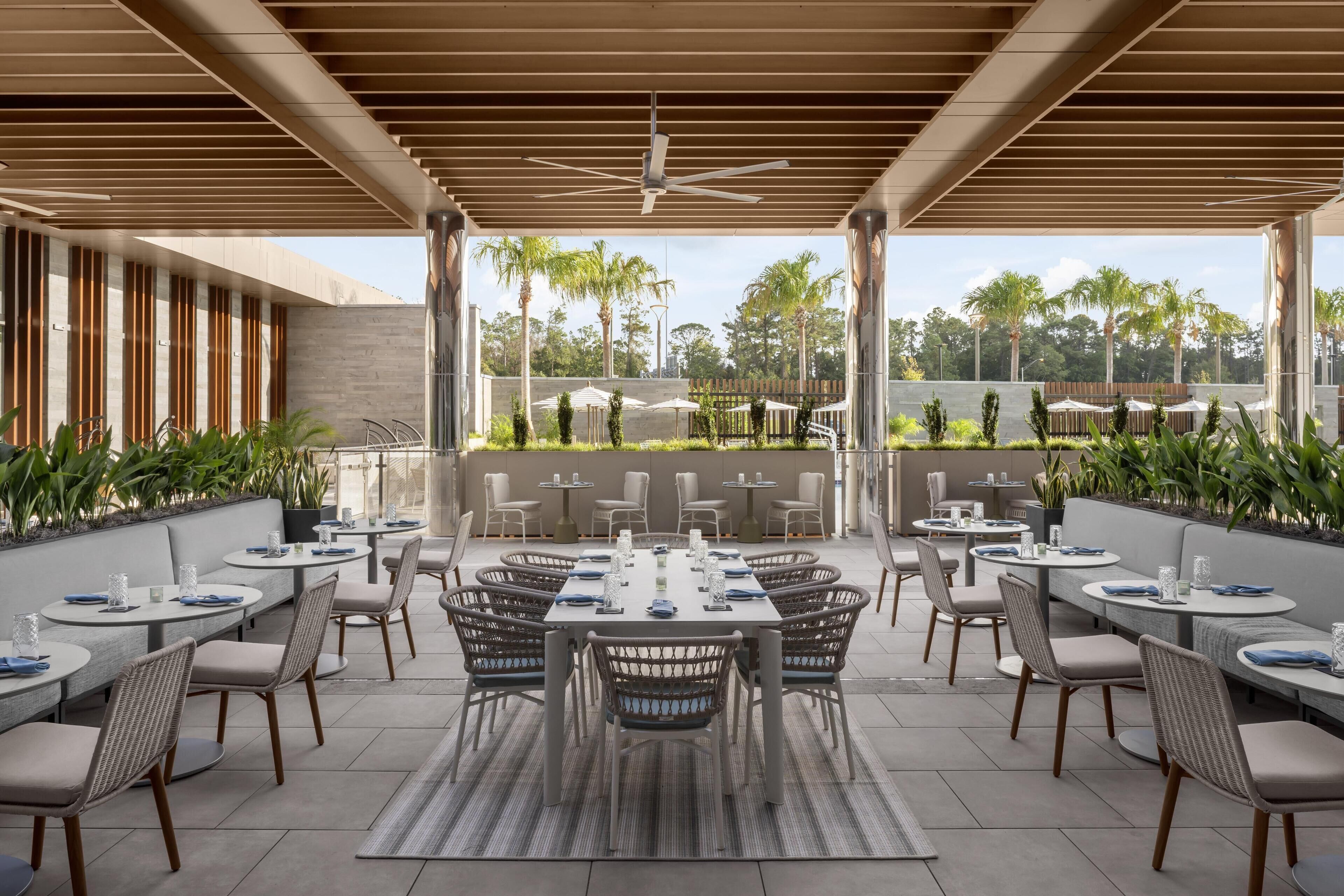 Un espacio de comedor al aire libre con techo, mesas y sillas, un ventilador de techo y vistas a palmeras y follaje.