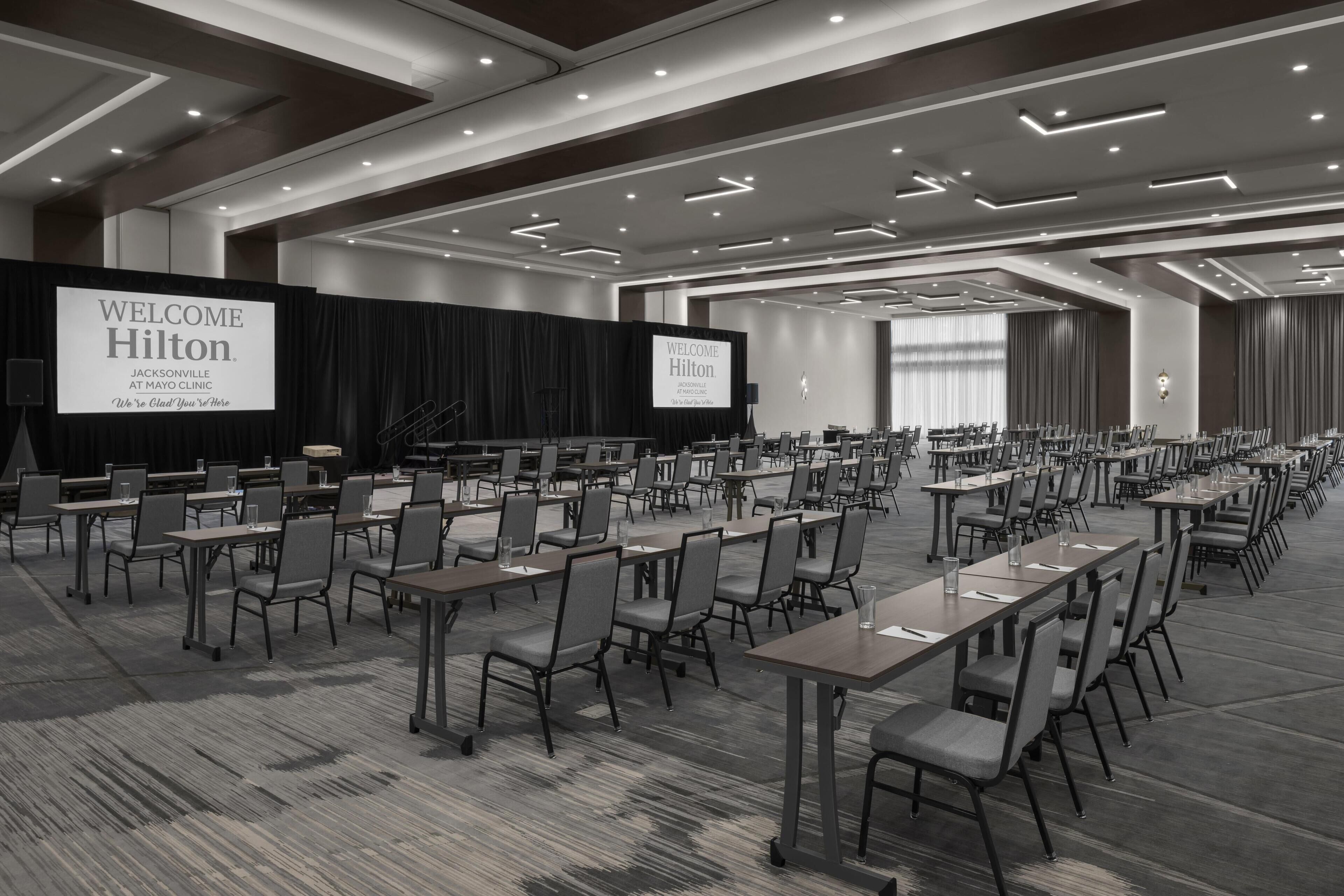 Une grande salle de conférence avec de nombreuses tables et chaises disposées en rangées, une scène ornée d’une banderole « Bienvenue chez Hilton », et un design de plafond moderne.