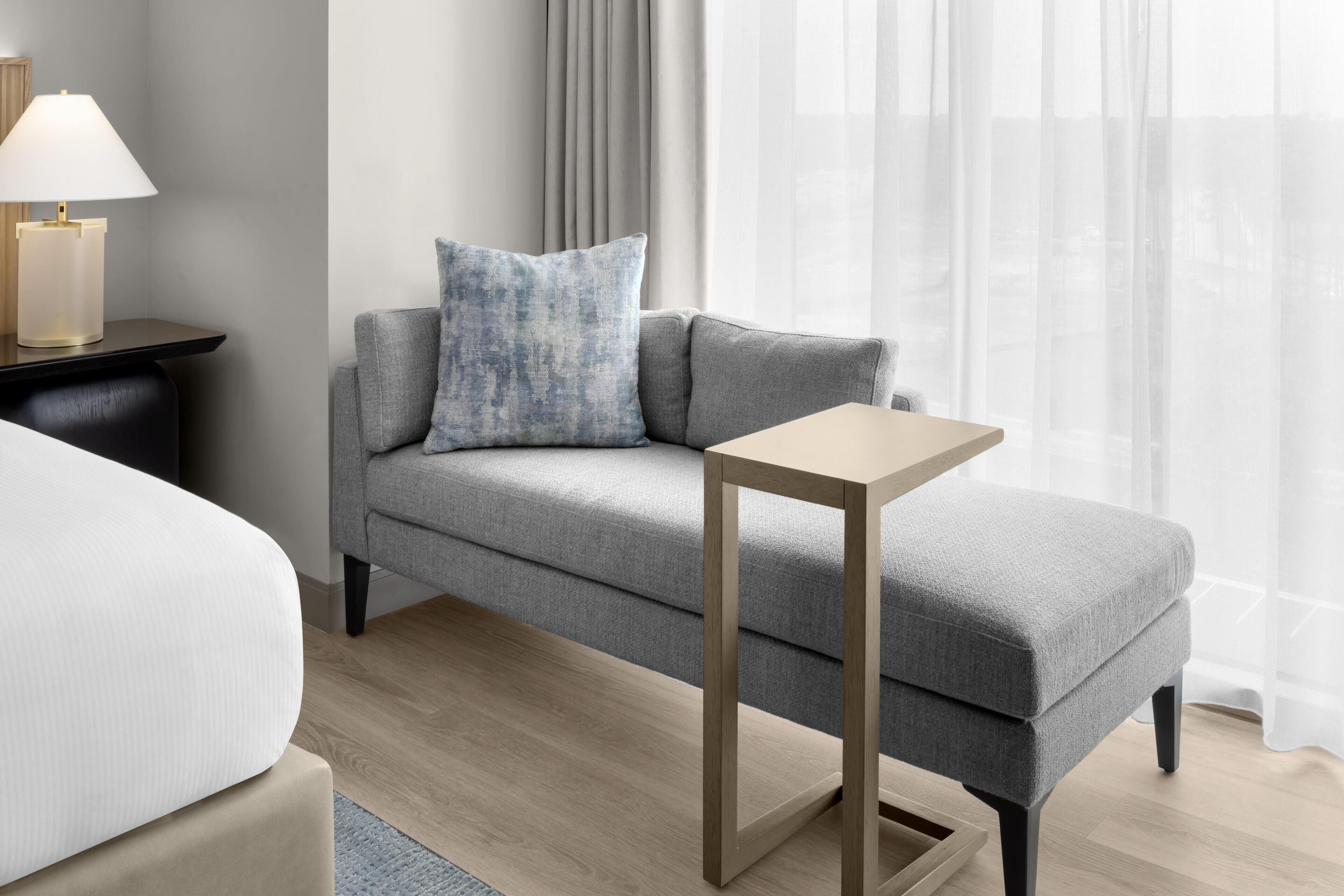 A modern hotel room with a grey sofa, a small wooden side table, and a lamp.