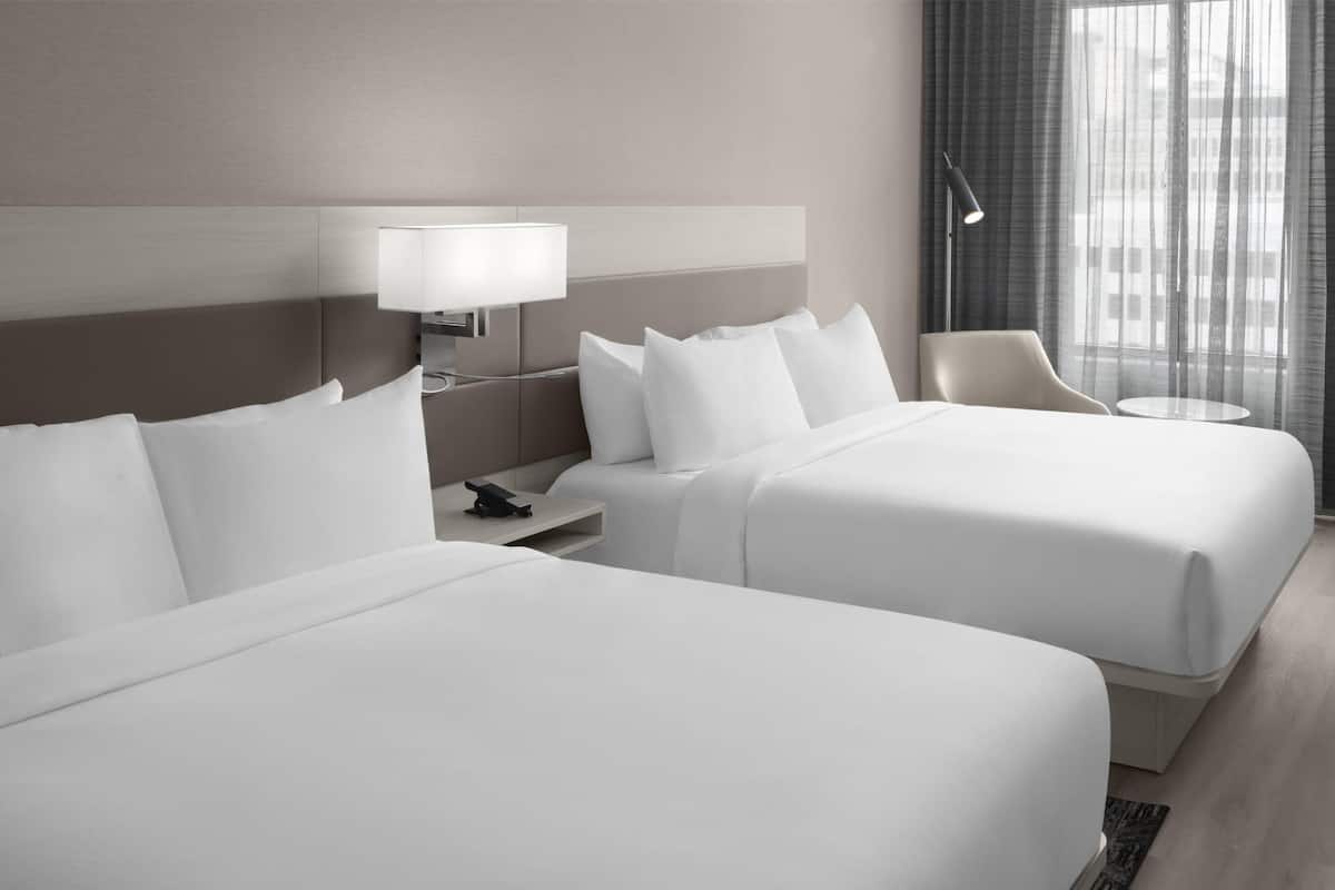 A hotel room with two beds, a bedside table with a lamp, a chair, and a view of the outside through a window with curtains.